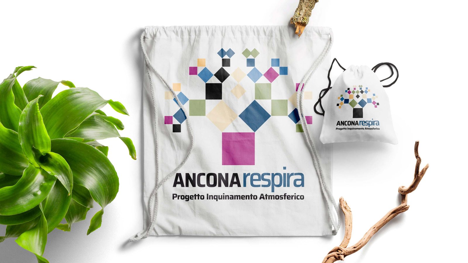 Due sacche di cotone bianco su fondo chiaro, stampate con il marchio Ancona Respira: un albero fatto di quadrati colorati, e sotto la scritta Progetto Inquinamento Atmosferico. Accanto una pianta verde e due rami secchi.