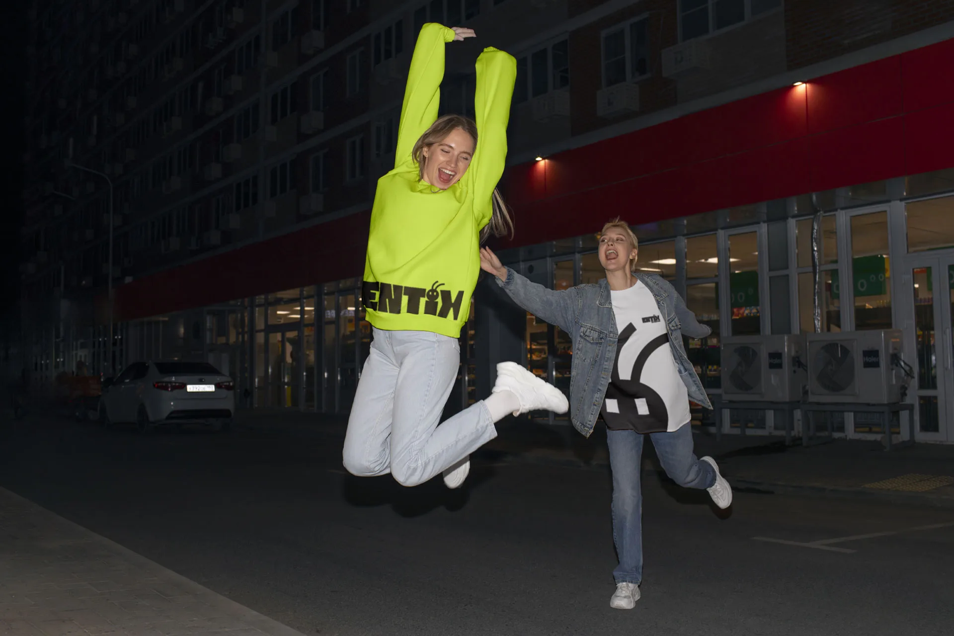 Due donne che saltano e ridono di notte in strada, una con felpa verde acido Entix, l'altra con maglietta bianca e nera Entix sotto una giacca di jeans.