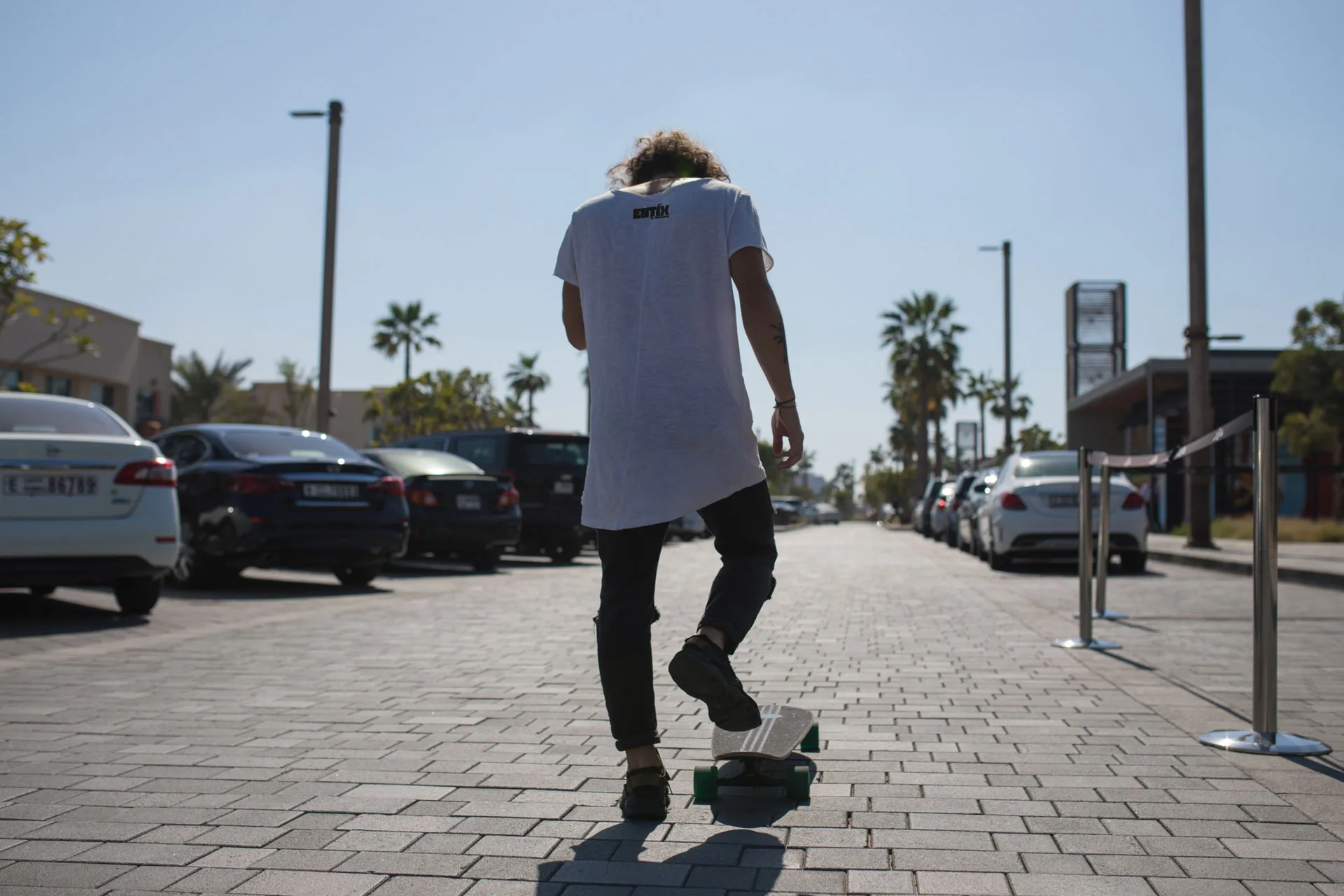 Uomo di spalle con maglietta bianca e piccolo logo Entix, un piede sullo skateboard, su una strada pedonale con palme e auto parcheggiate.
