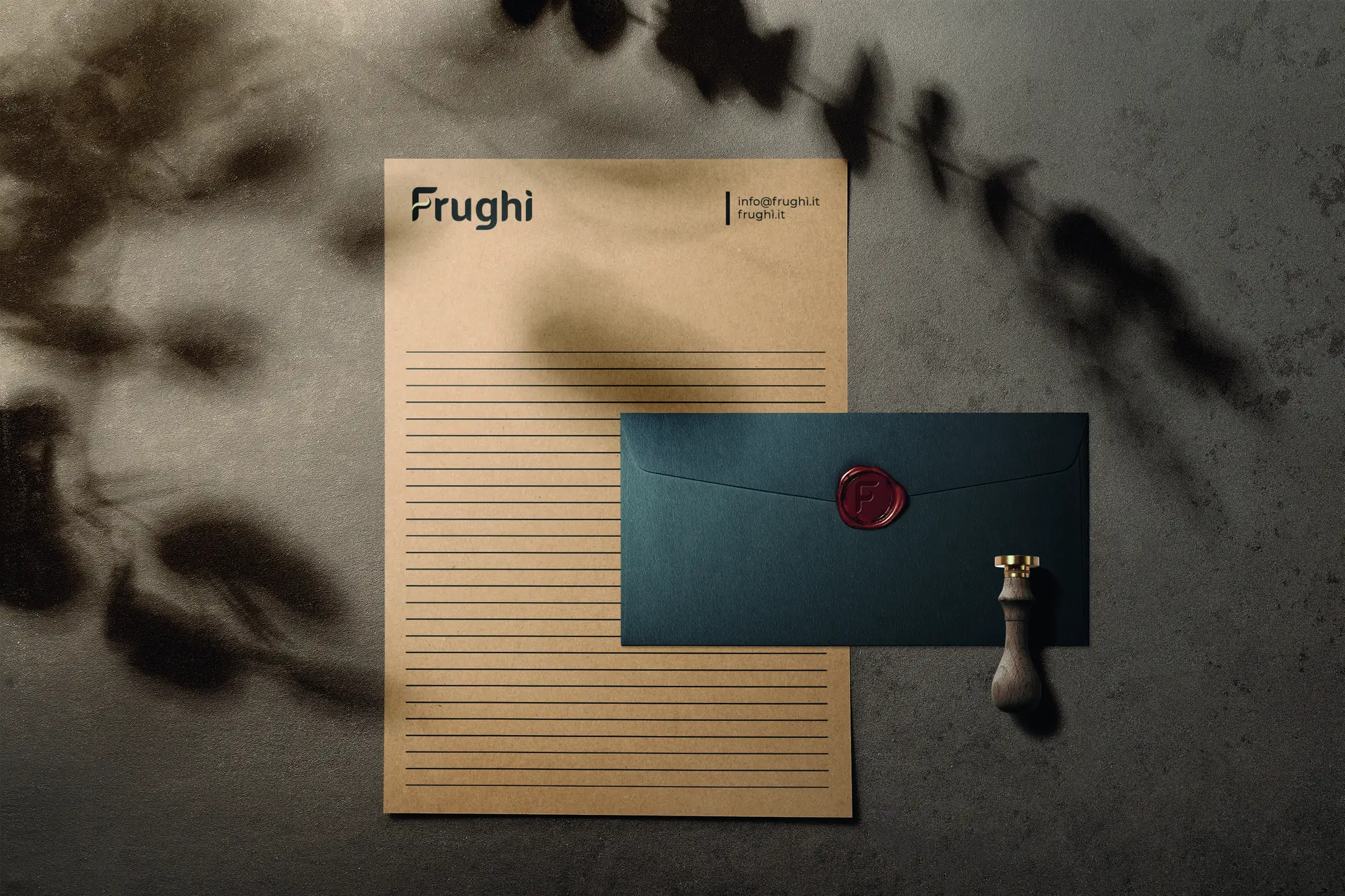 Carta intestata in Kraft rigata e busta blu scuro chiusa con un bollo di ceralacca rossa impresso con la F, fotografate dall'alto su una superficie scura con l'ombra di un ramo.