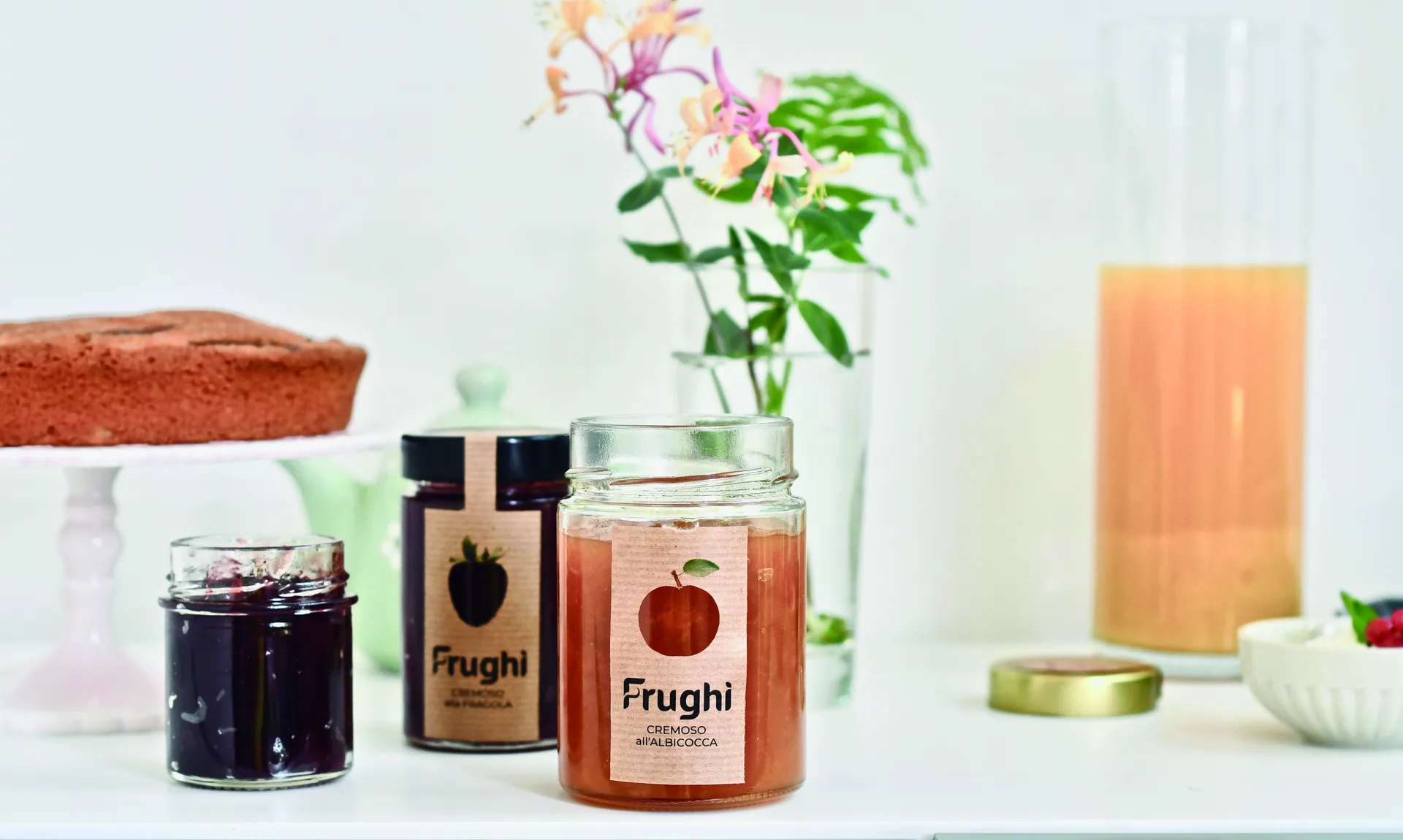 Tre vasetti Frughì su un piano bianco accanto a una torta: etichette in carta Kraft con la sagoma del frutto ritagliata, il nome in nero e sotto il gusto — cremoso alla fragola, cremoso all'albicocca.