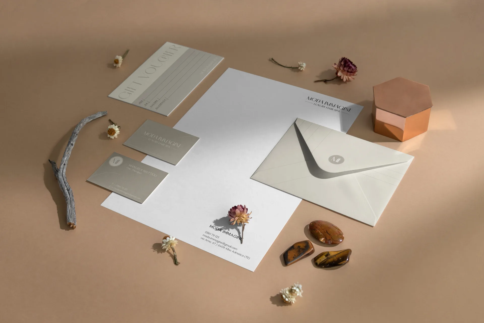 La coordinata vista dall'alto su fondo cipria: gift voucher, carta intestata, due biglietti da visita verde salvia, busta avorio con il monogramma tondo sul lembo, fiori secchi e pietre attorno.