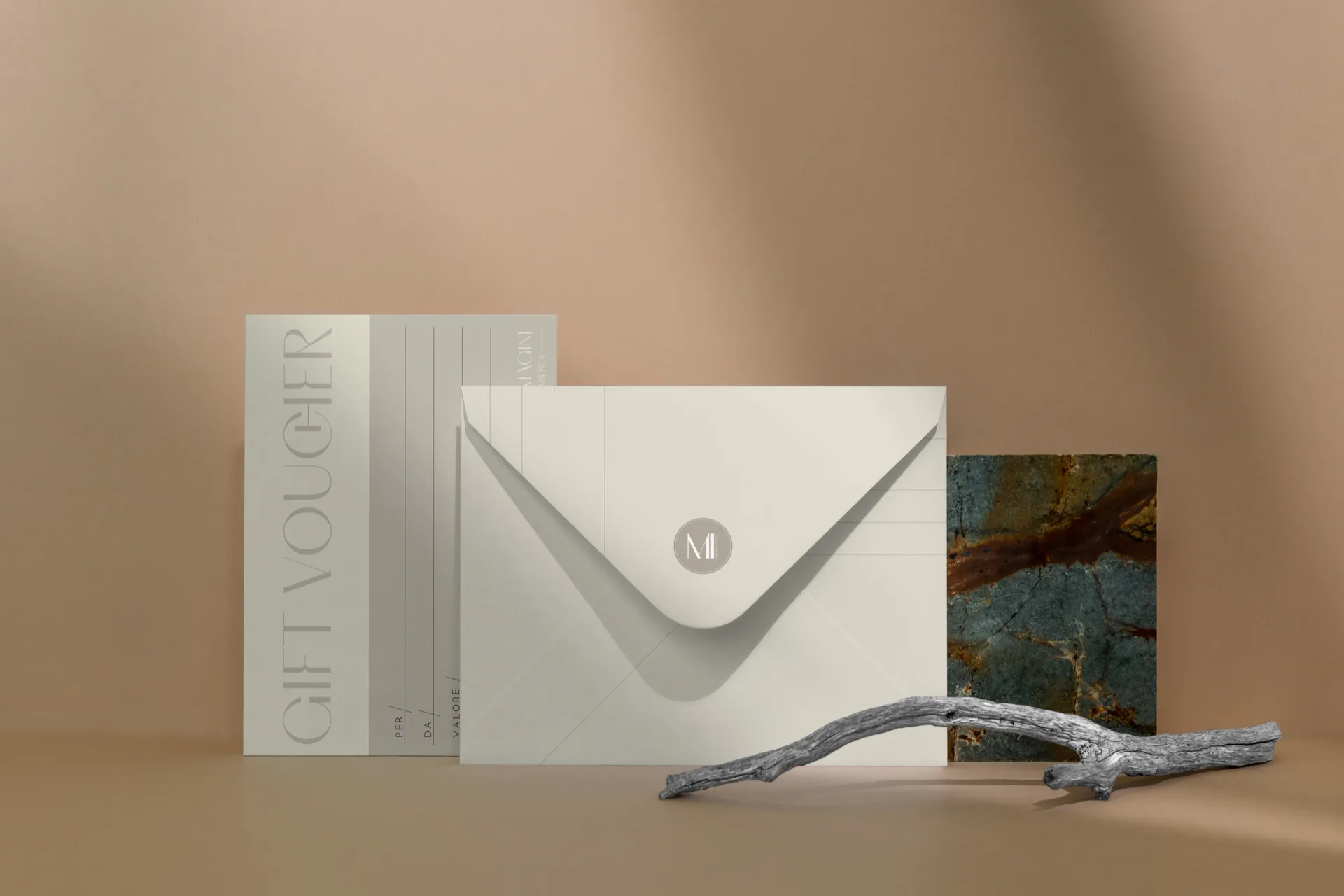 Gift voucher in piedi contro un fondo cipria, con la scritta GIFT VOUCHER in verticale e le righe da compilare, accanto a una busta avorio chiusa dal monogramma tondo.