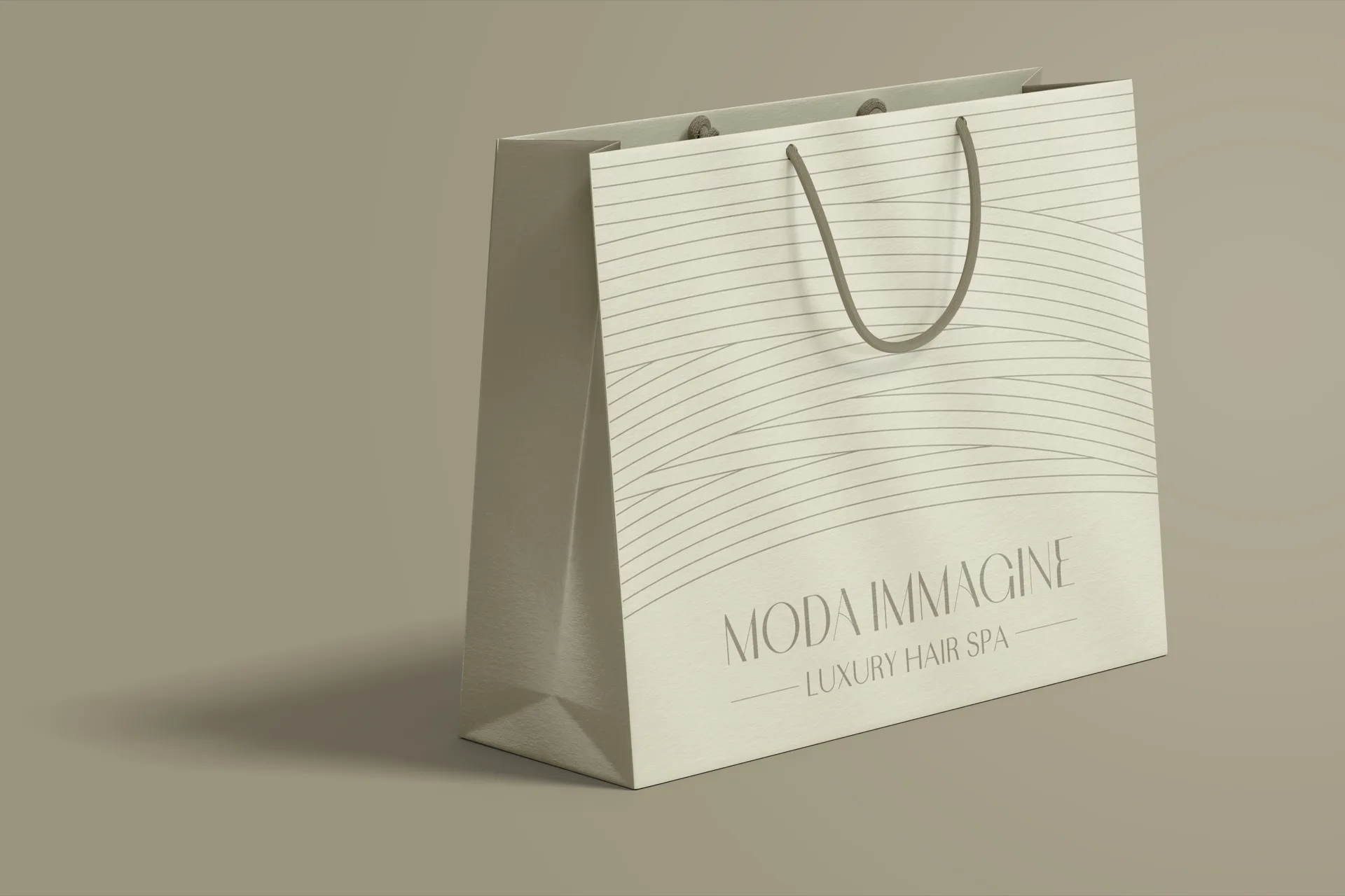 Shopper grande in carta avorio con manici in corda, di tre quarti su fondo verde oliva: linee sottili che ondeggiano nella metà alta e sotto il nome per esteso.