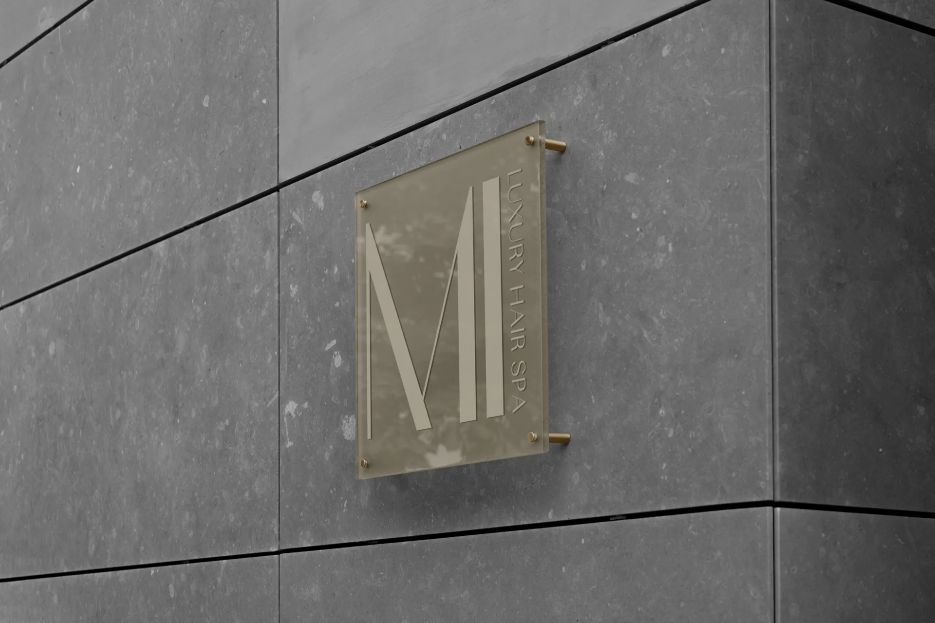 Targa in plexiglas fumé fissata a una parete in pietra grigia con quattro distanziali in ottone: dentro, il monogramma con la M e la I e la dicitura in verticale.