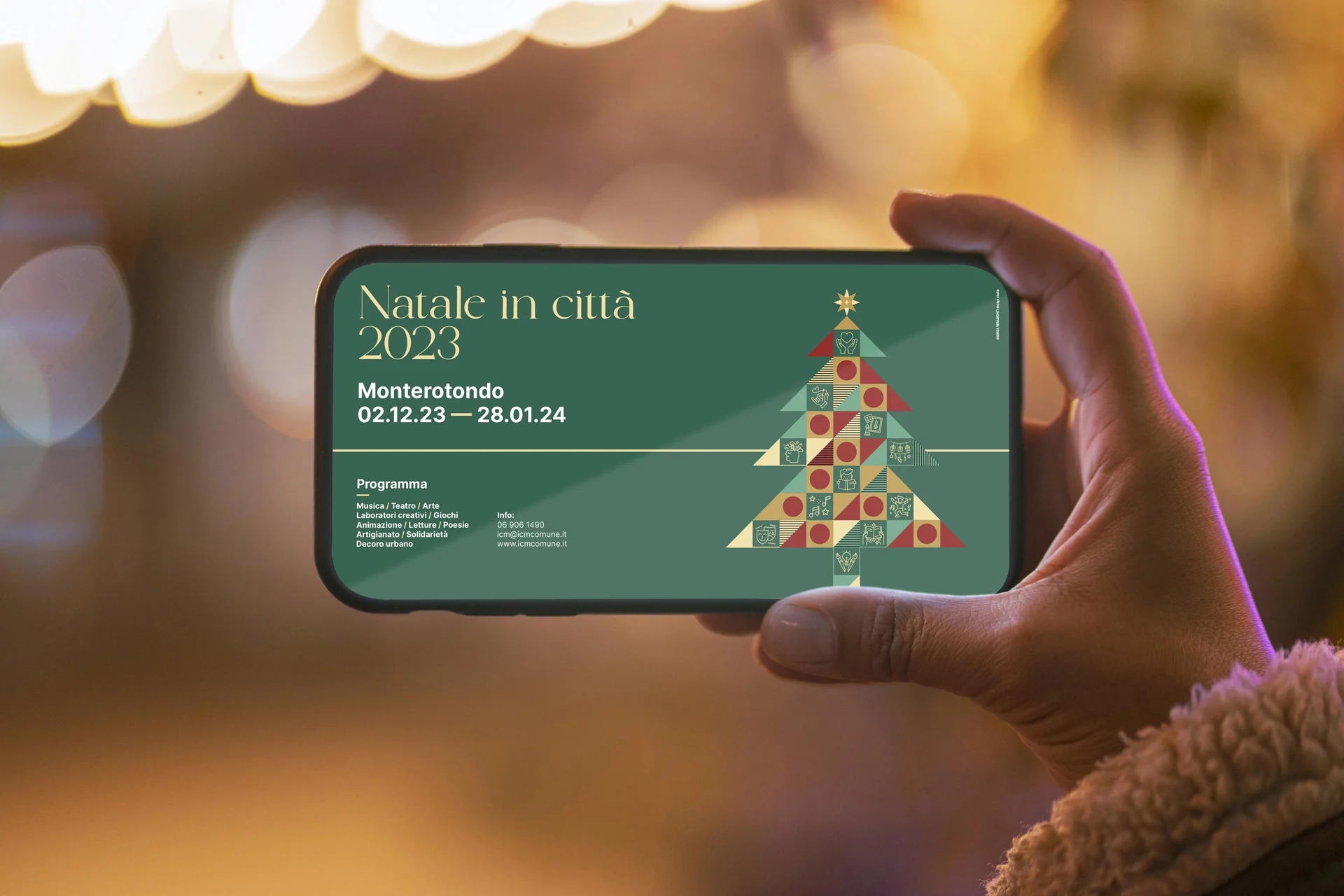Telefono tenuto in orizzontale davanti alle luminarie di una via: sullo schermo la testata del programma digitale, con titolo, date e l'albero sulla destra.