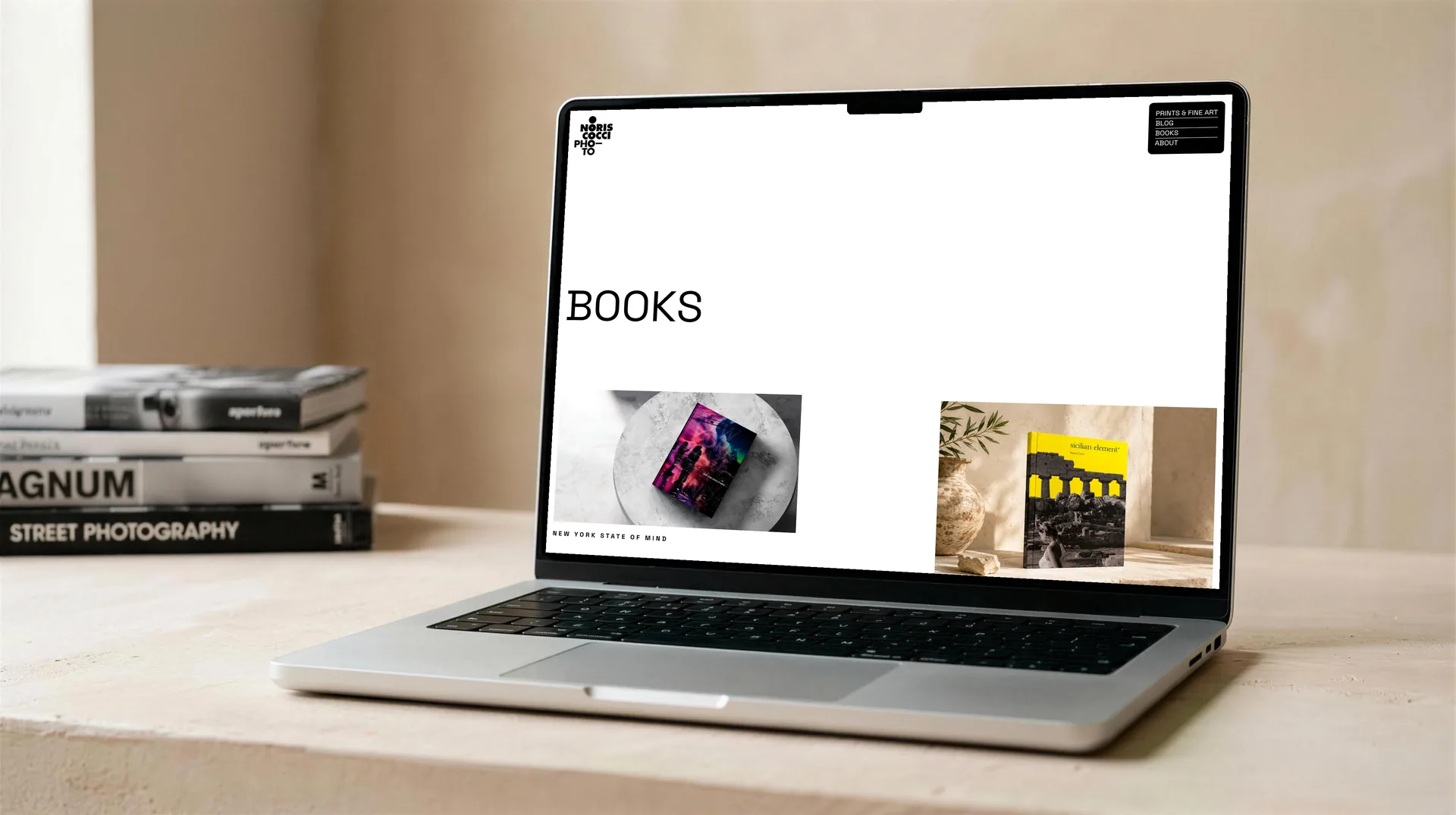 Mock-up: la sezione Books sullo schermo di un portatile aperto su un piano di calce chiara, con il titolo a sinistra e due copertine di libri fotografate in scena; accanto al portatile una pila di libri di fotografia.