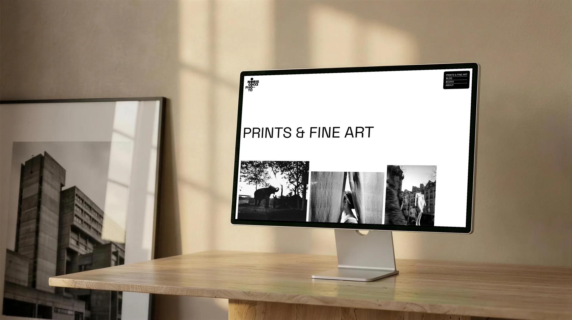 Mock-up: la sezione Prints & Fine Art di noriscocci.com su un monitor appoggiato a una scrivania di legno chiaro, con il titolo grande a sinistra e sotto tre fotografie in bianco e nero a quote diverse; a lato una stampa incorniciata appoggiata al muro.