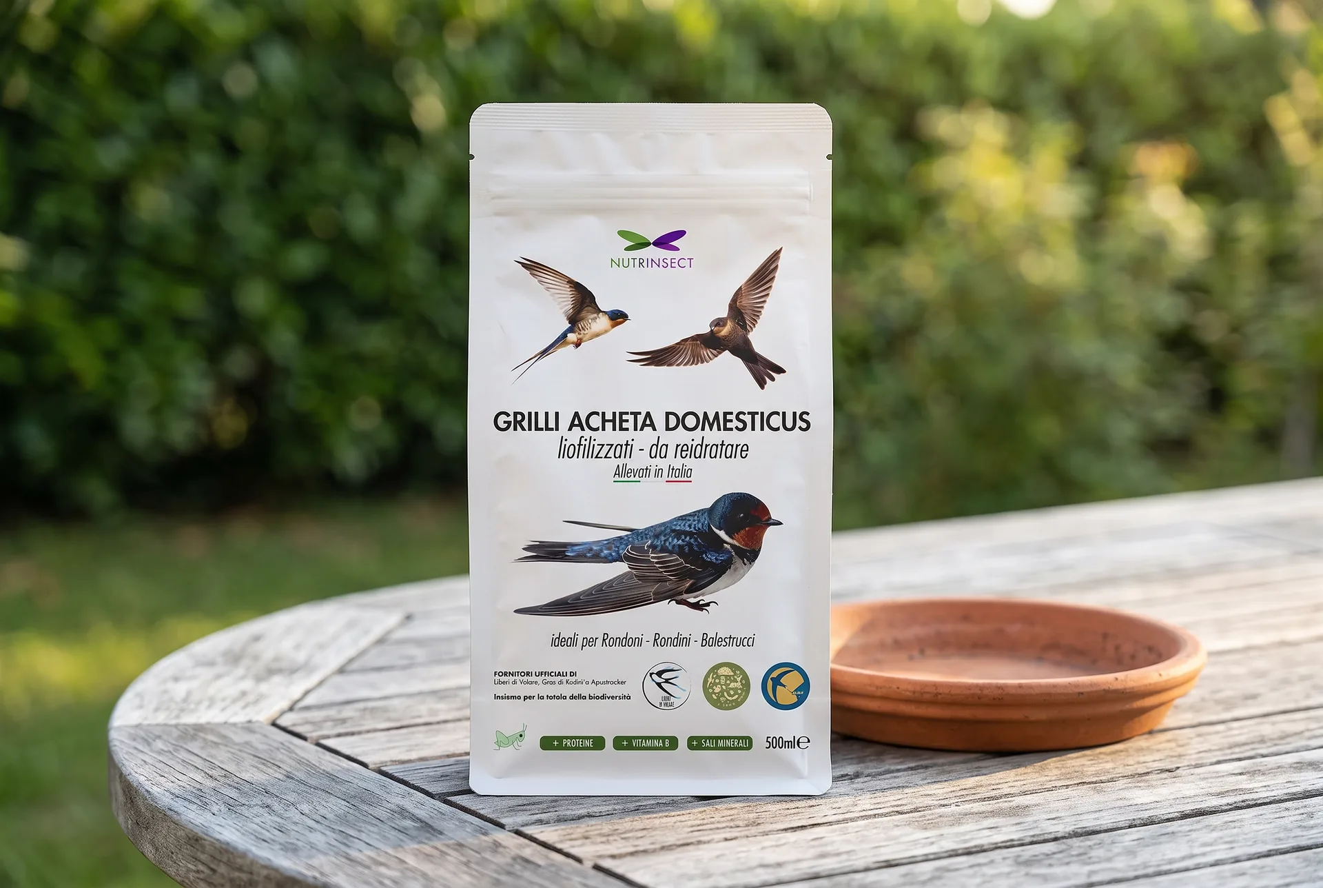 Mock-up generato: la busta dei grilli liofilizzati per rondoni e balestrucci in piedi su un tavolo da giardino di legno grigio, con un sottovaso di terracotta accanto e una siepe fuori fuoco dietro.