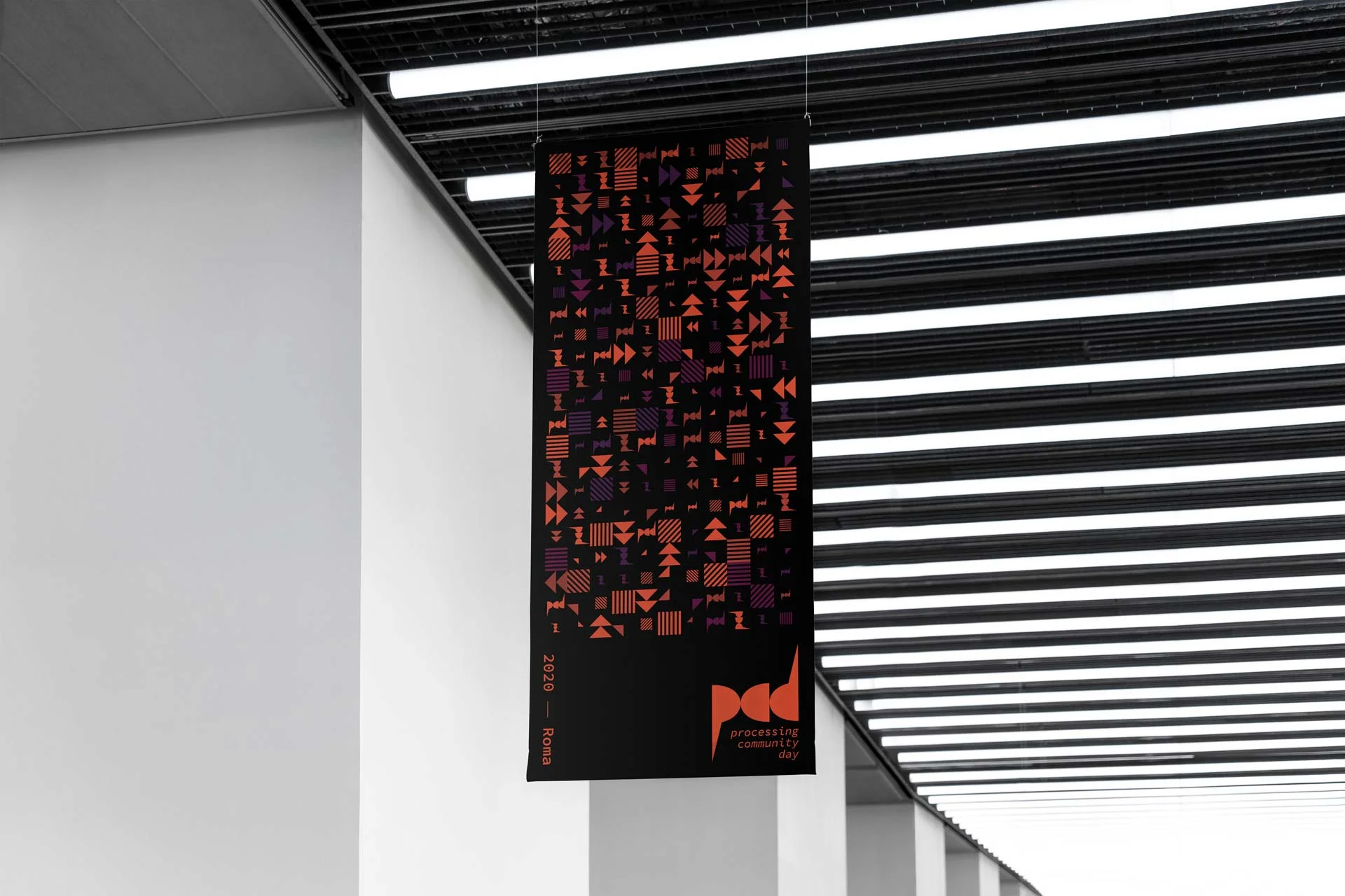 Bandiera verticale nera appesa al soffitto di un corridoio industriale, coperta di elementi geometrici arancioni e viola, con il marchio pcd in basso a destra e la scritta verticale 2020 — Roma sul lato sinistro.