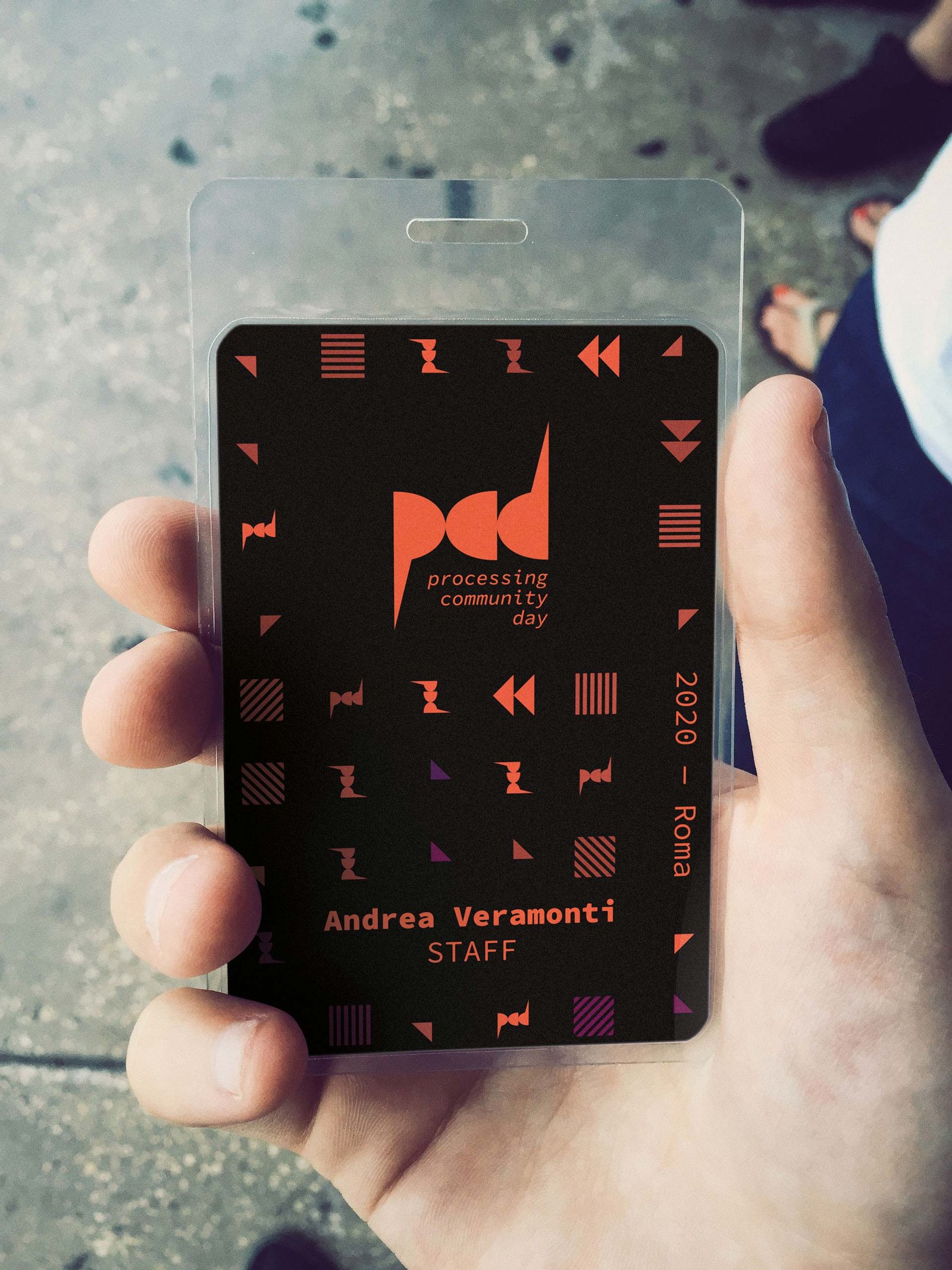 Mano che tiene il pass dell'evento in una custodia trasparente: cartoncino nero con il marchio pcd al centro, il pattern intorno, la scritta verticale 2020 — Roma e in basso il nome Andrea Veramonti con la qualifica STAFF.