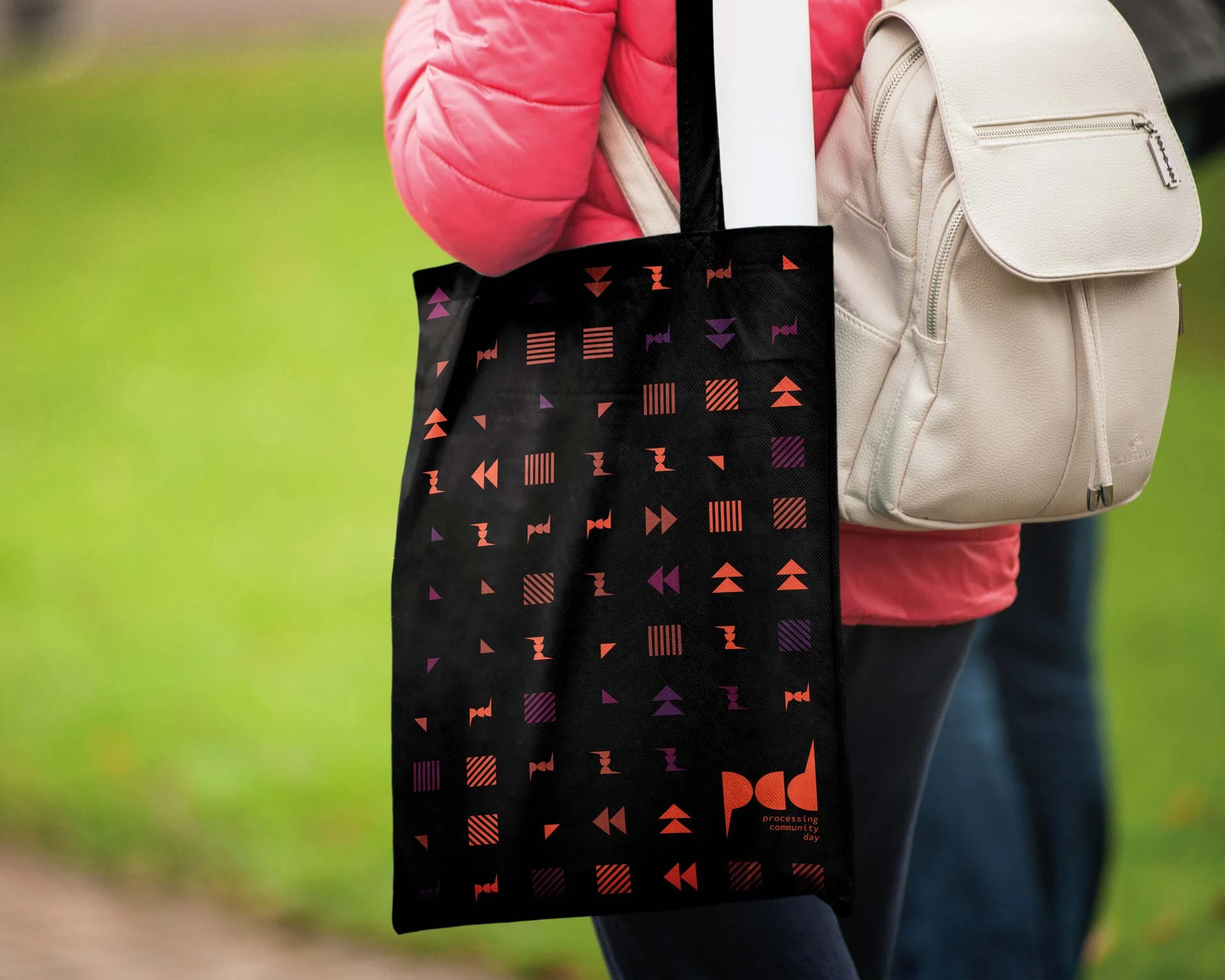 Persona con giacca rosa che porta a spalla una shopper nera stampata con il pattern e il marchio pcd in arancione, su uno sfondo di prato verde.