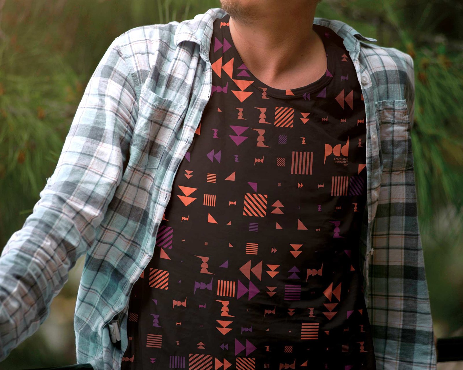 Uomo con camicia a quadri aperta su una maglietta nera interamente coperta dal pattern in arancione e viola, con il marchio pcd all'altezza del petto.