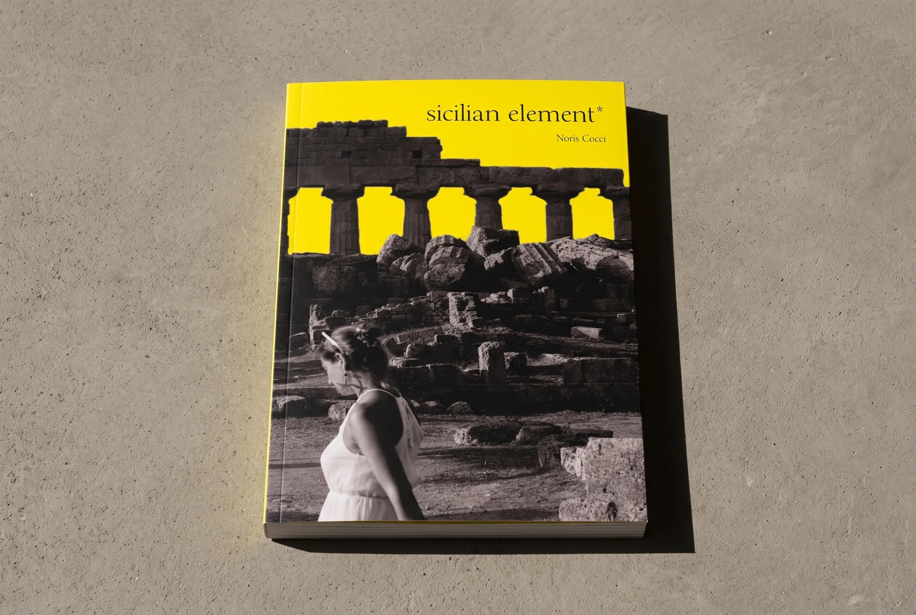 Mock-up del libro chiuso, appoggiato su un piano di cemento chiaro e ripreso da poco sopra: copertina gialla, fotografia in bianco e nero di un tempio greco e di una ragazza vestita di bianco, titolo in minuscolo sul giallo.