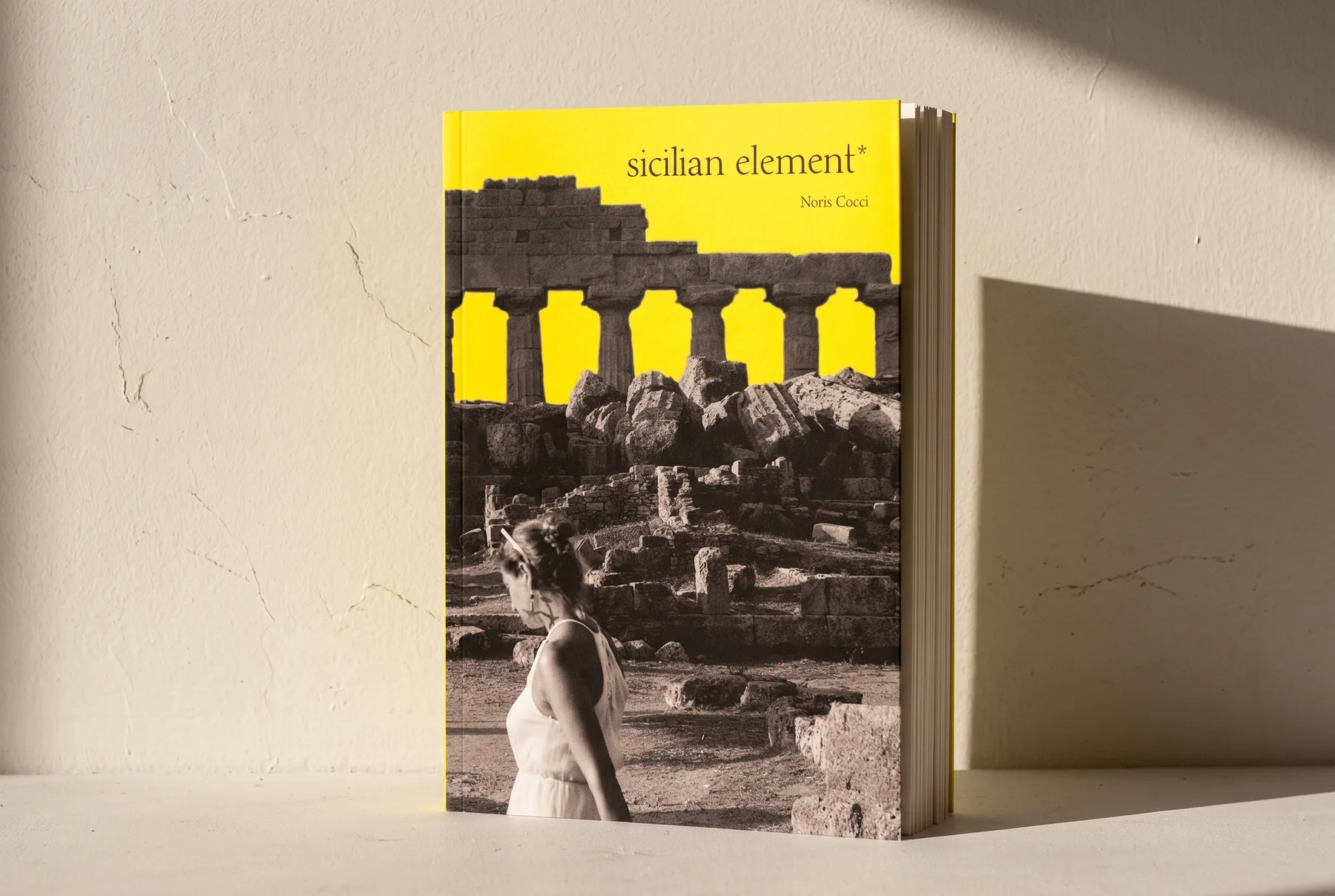 Mock-up del libro in piedi, appoggiato a un muro di intonaco chiaro con il sole di taglio che allunga l'ombra: si vede lo spessore della carta sul taglio anteriore.
