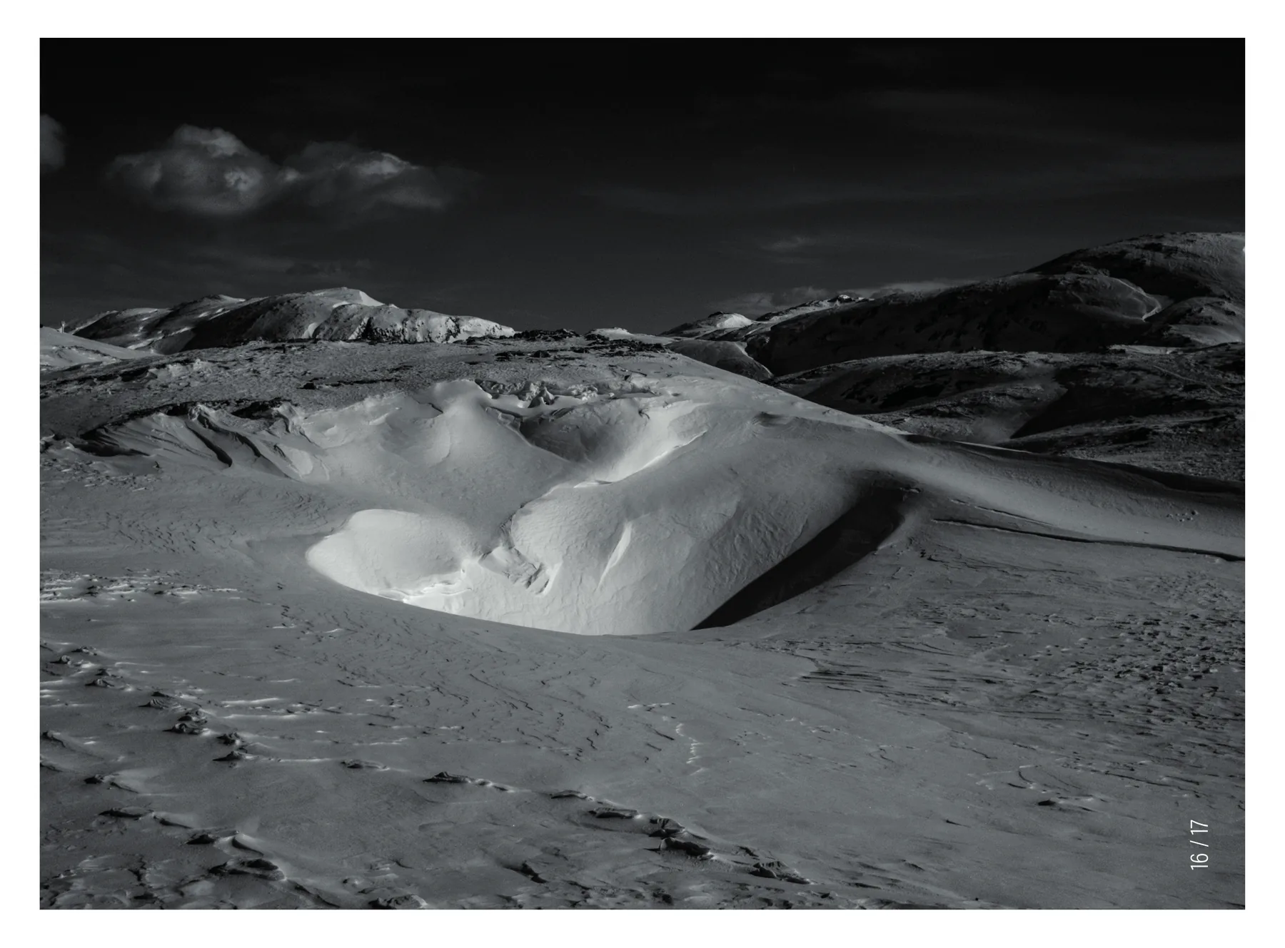 Doppia pagina 16 / 17: una distesa di neve ventata sotto un cielo nero, con le dune scavate dal vento e le impronte in primo piano, stampata scura da un bordo all'altro.