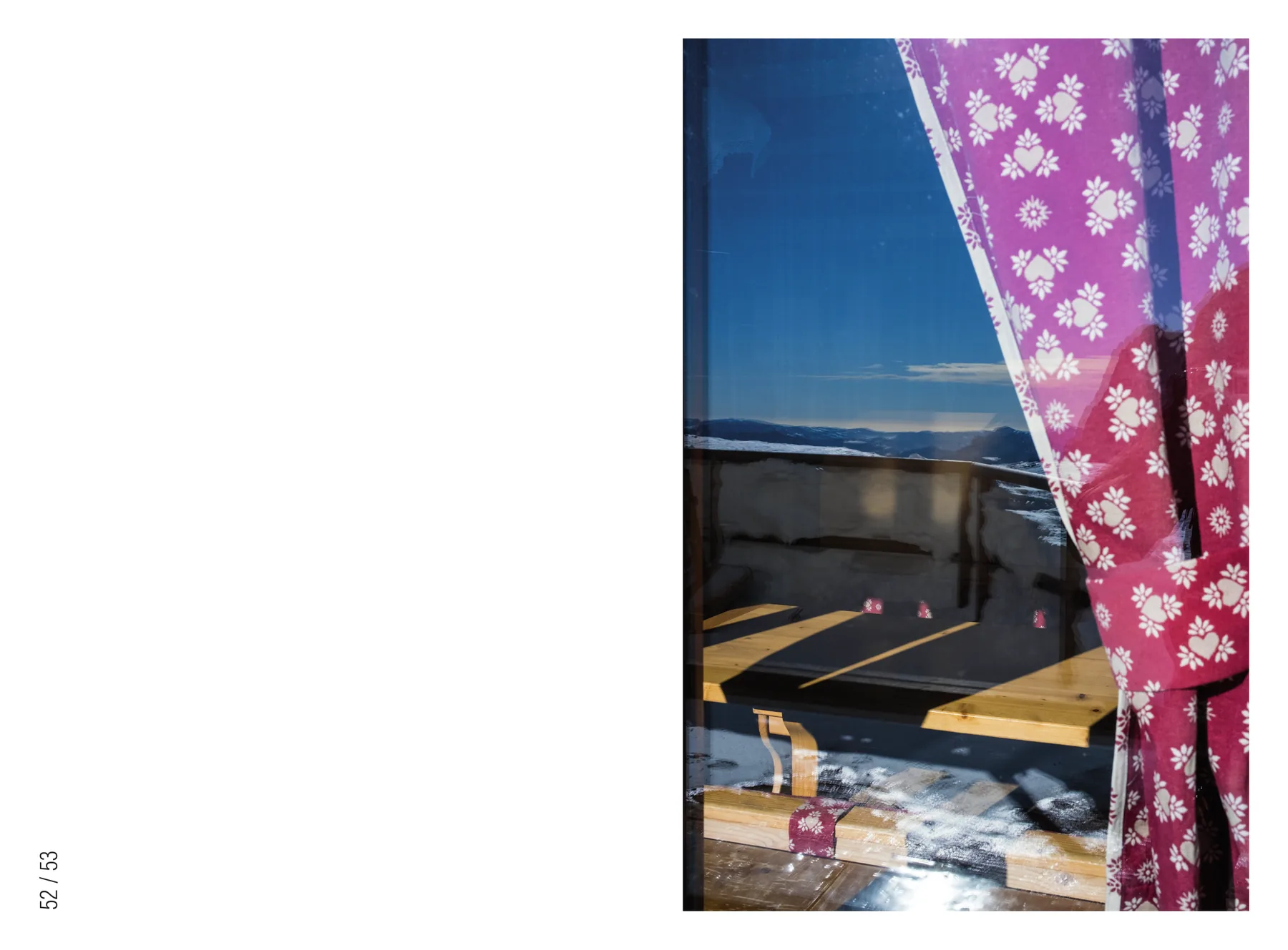 Doppia pagina con la pagina di sinistra vuota e a destra una fotografia verticale: una tenda a fiori rosa scostata su una finestra, oltre la quale si vede la neve e i tavoli di legno.