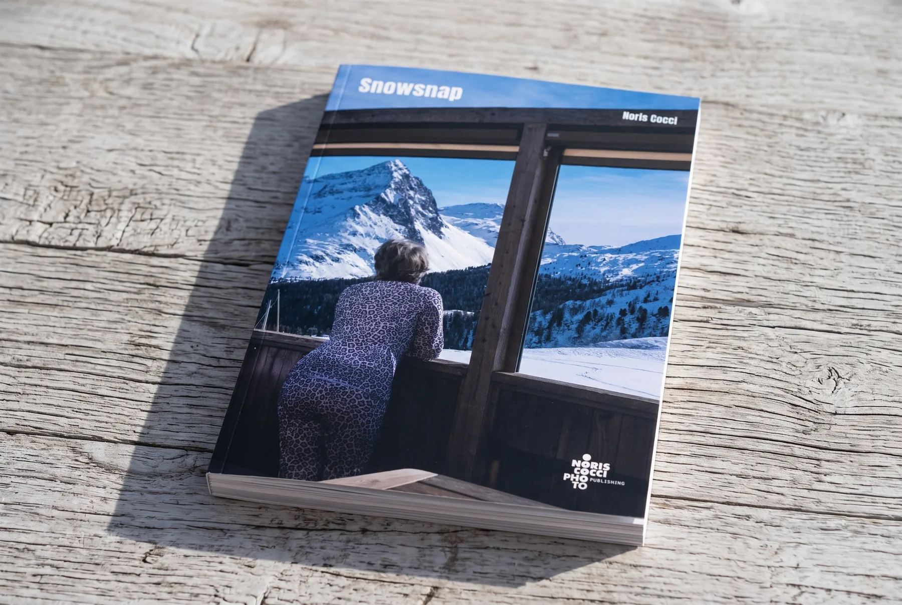 Mock-up del libro chiuso su un tavolo di legno chiaro, ripreso da poco sopra: in copertina la veduta presa da dentro un rifugio, con le vetrate che inquadrano le cime innevate, e il titolo Snowsnap in bianco.