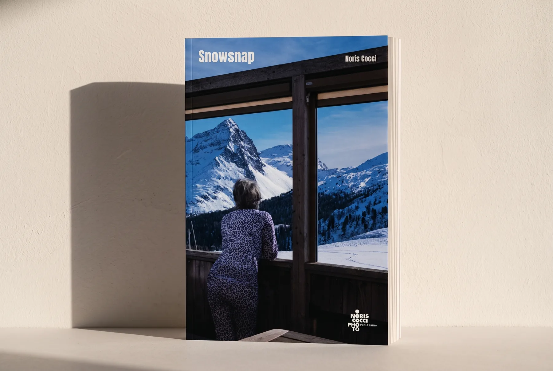 Mock-up del libro in piedi contro un muro di intonaco chiaro, con il sole basso di taglio che allunga l'ombra e mette in luce lo spessore della carta.