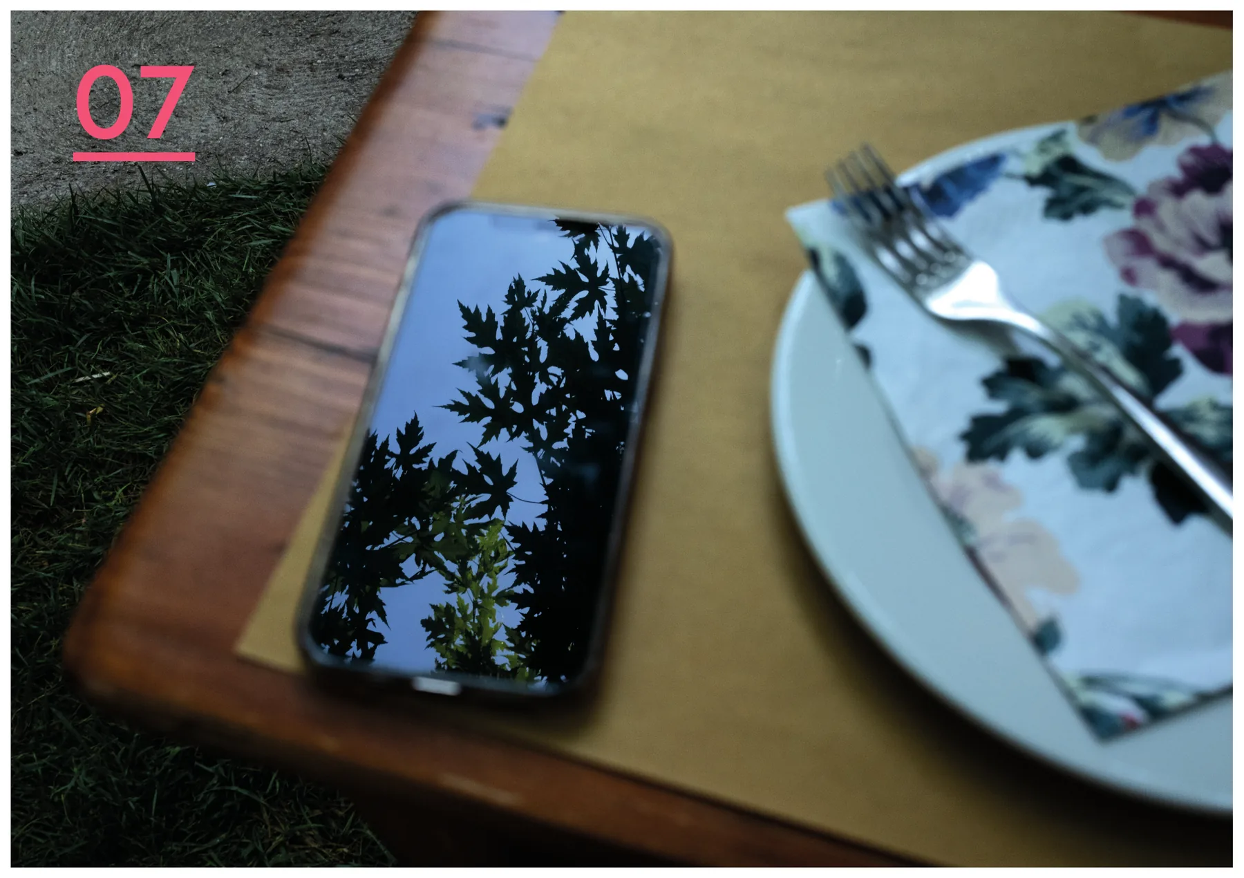 Doppia pagina a piena immagine: un tavolo di legno all'aperto con un telefono che riflette i rami di un albero e, accanto, un piatto a fiori con le posate; il numero 07 sottolineato in alto a sinistra.