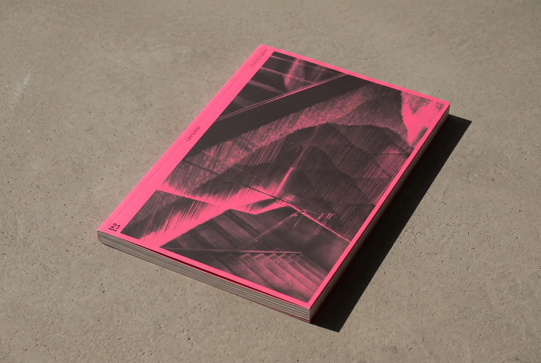 Mock-up del libro chiuso su un piano di cemento chiaro, ripreso da poco sopra: copertina rosa fluorescente con dentro una fotografia in duotone nero su rosa e il numero 21 sottolineato in basso a sinistra.
