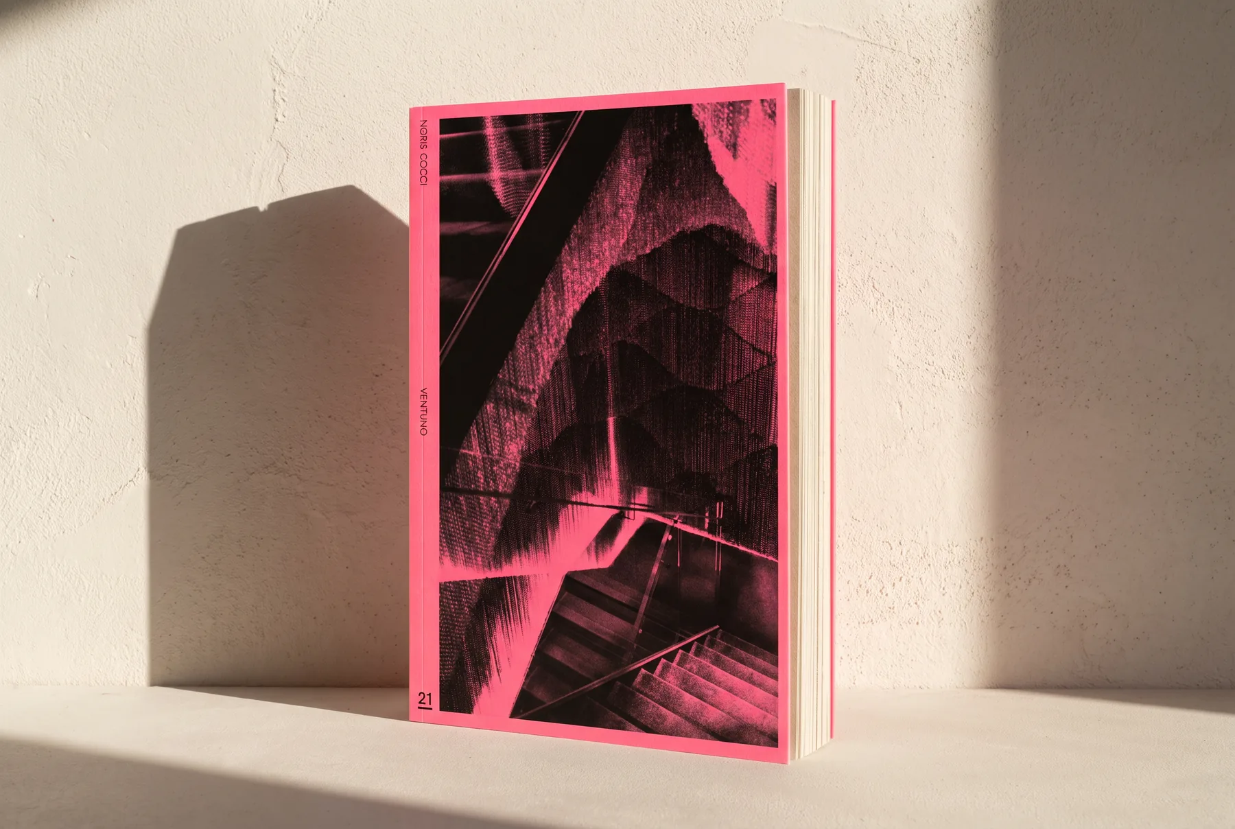Mock-up del libro in piedi contro un muro di intonaco chiaro, col sole di taglio che allunga l'ombra: si vede il taglio anteriore e lo spessore delle pagine.