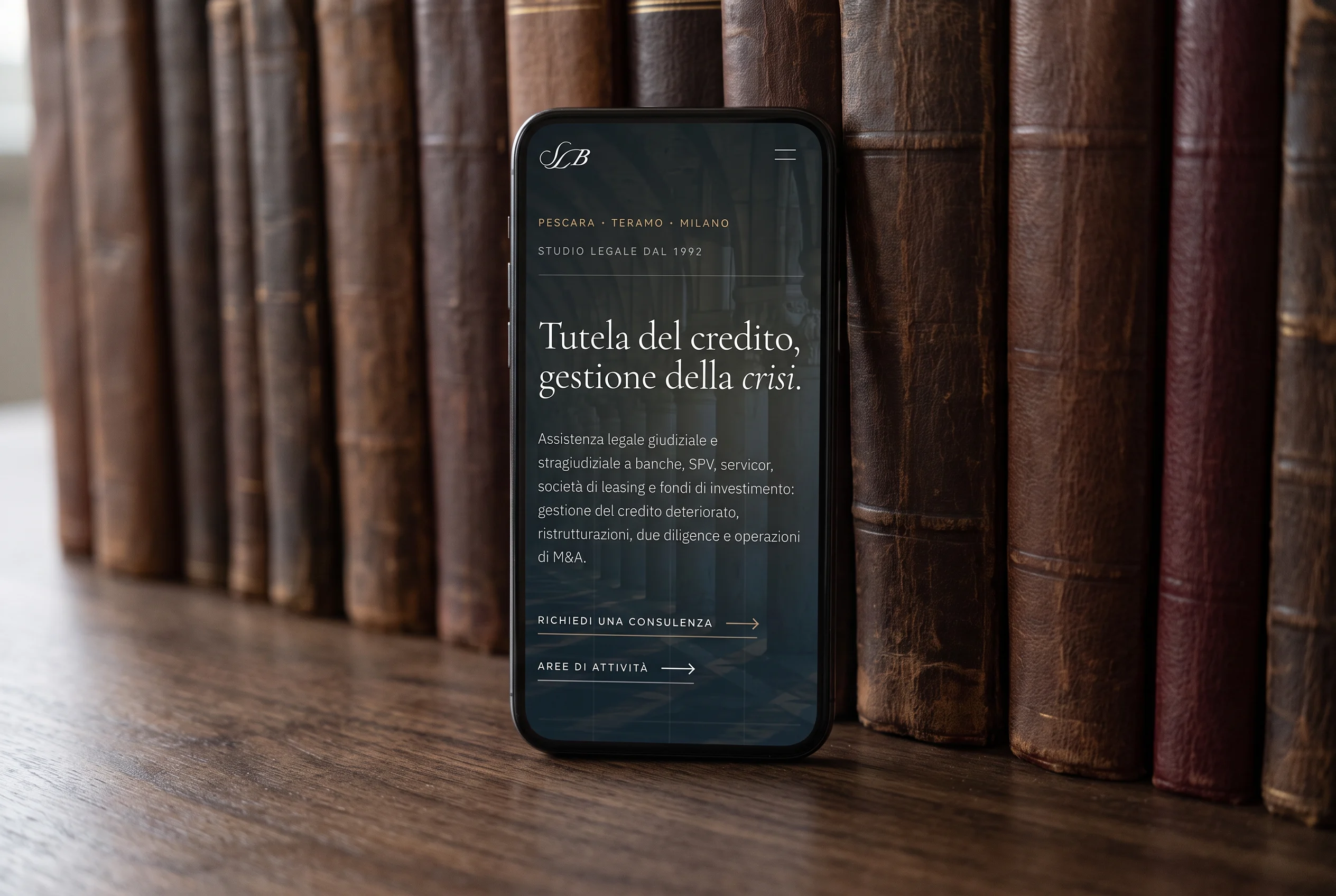 Mock-up: la home del sito sul telefono, appoggiato in verticale a una fila di vecchi libri di legge; il titolo e il testo introduttivo vanno a capo su una colonna sola e il menu diventa un’icona in alto a destra.